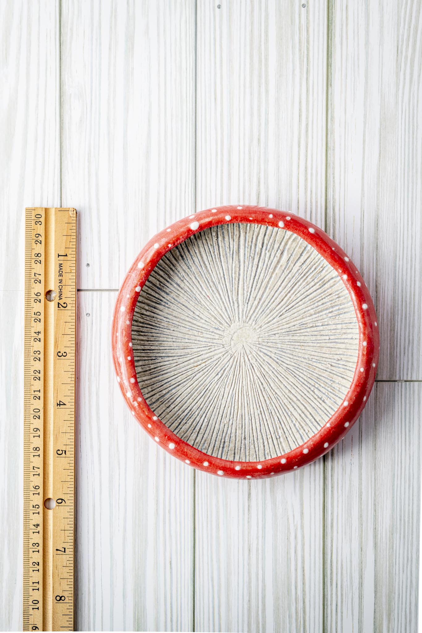 Decorative small plate with red rim and white center on a wooden surface, accompanied by a ruler for scale. Handmade pottery by TJ